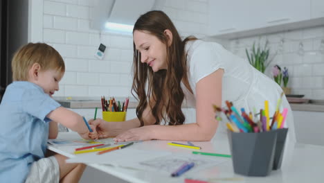 Young-beautiful-mom-and-son-draw-with-colored-pencils-sitting-at-the-table-in-the-kitchen